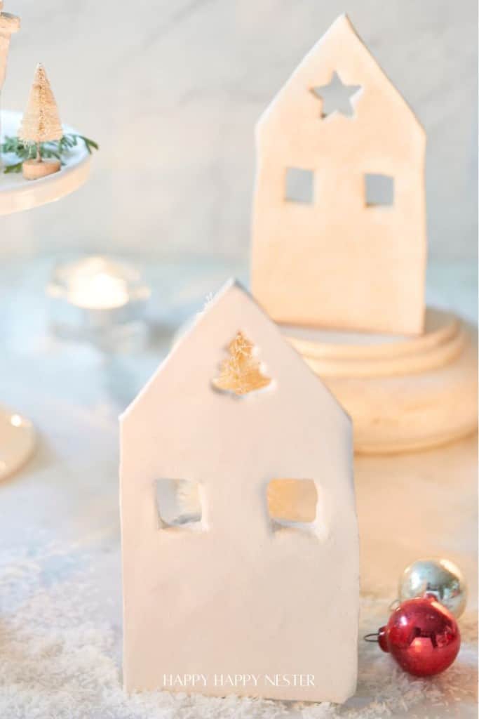 Two white, handmade clay house decorations with cut-out windows and shapes on top, set on a snowy surface—these pieces are my favorite Christmas decor, surrounded by festive ornaments and miniature trees in the background.