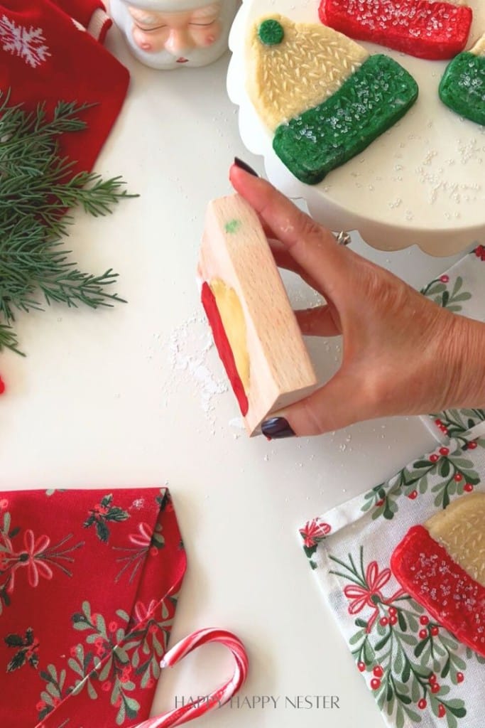 A hand holds a wooden stamp above a white table decorated with molded cookies recipe treats shaped like hats, festive napkins, a candy cane, evergreen sprigs, and a cake stand.