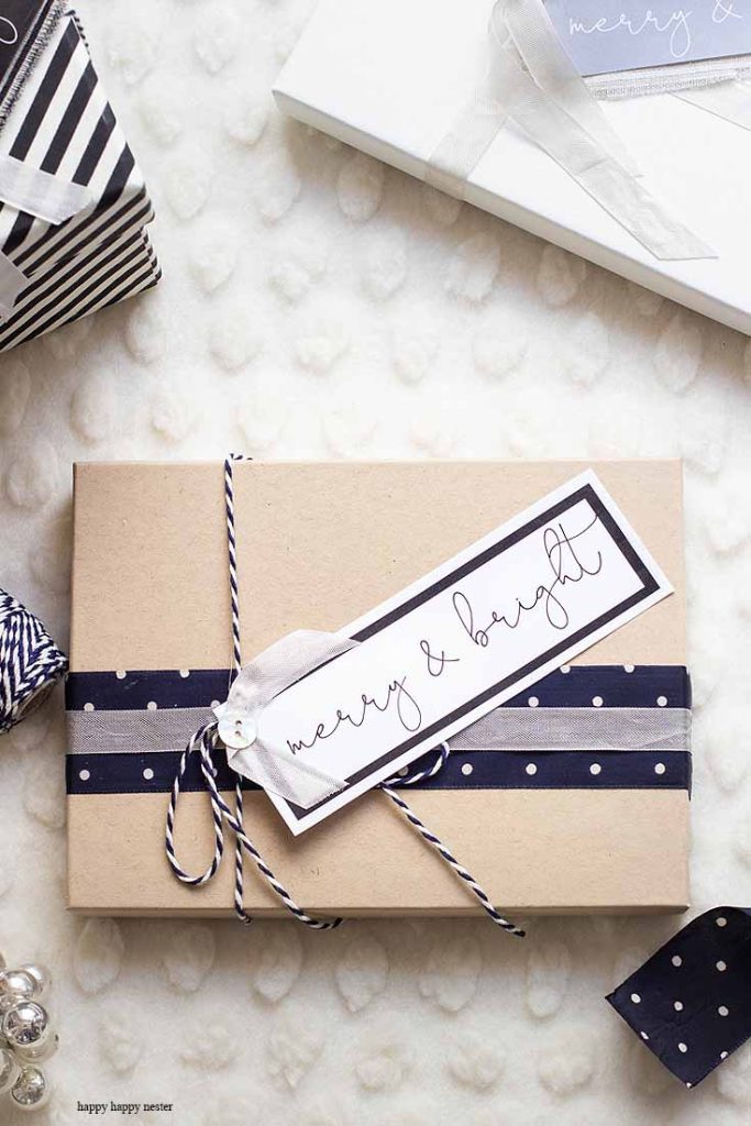 A gift wrapped in brown paper with navy and white polka dot ribbon, tied with a twine bow and a tag that reads “merry & bright.” Other presents and ribbon rolls are nearby on a textured white surface—my favorite gift wrap inspiration.