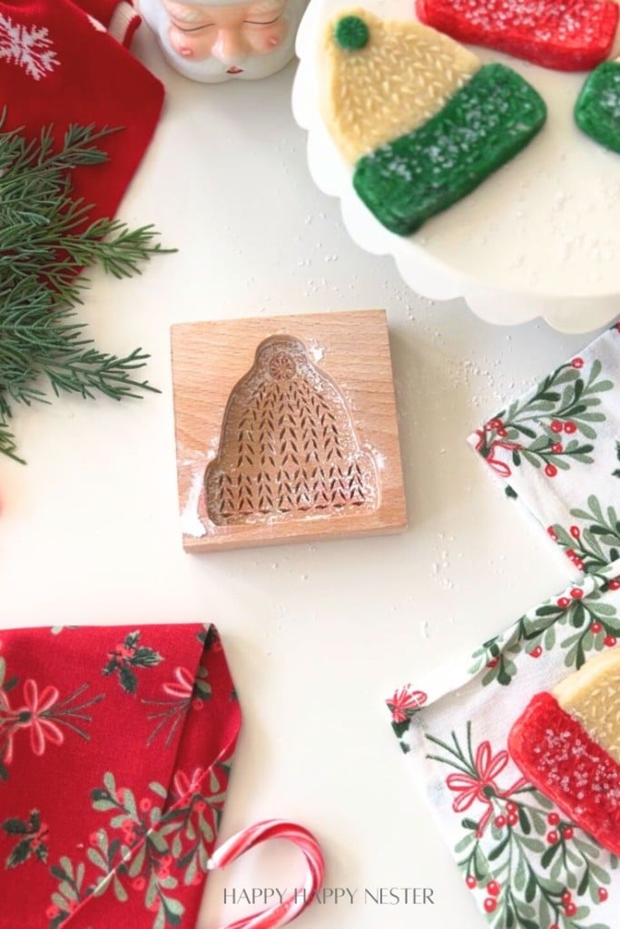 A wooden cookie mold shaped like a winter hat is placed on a white surface, surrounded by festive napkins, greenery, a candy cane, and cookies decorated as colorful winter hats.
