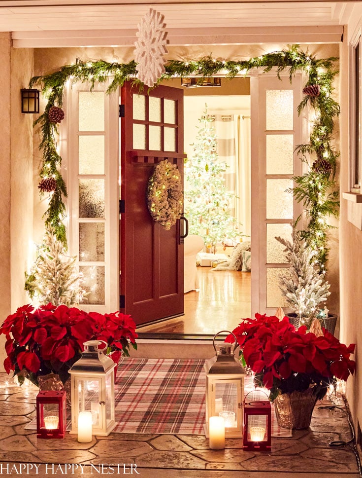 A festive front porch with a red door decorated with a wreath, surrounded by garland and pinecones—my favorite Christmas decor. Poinsettias, lanterns, candles, and small lit Christmas trees welcome you, while inside, a glowing tree can be seen.