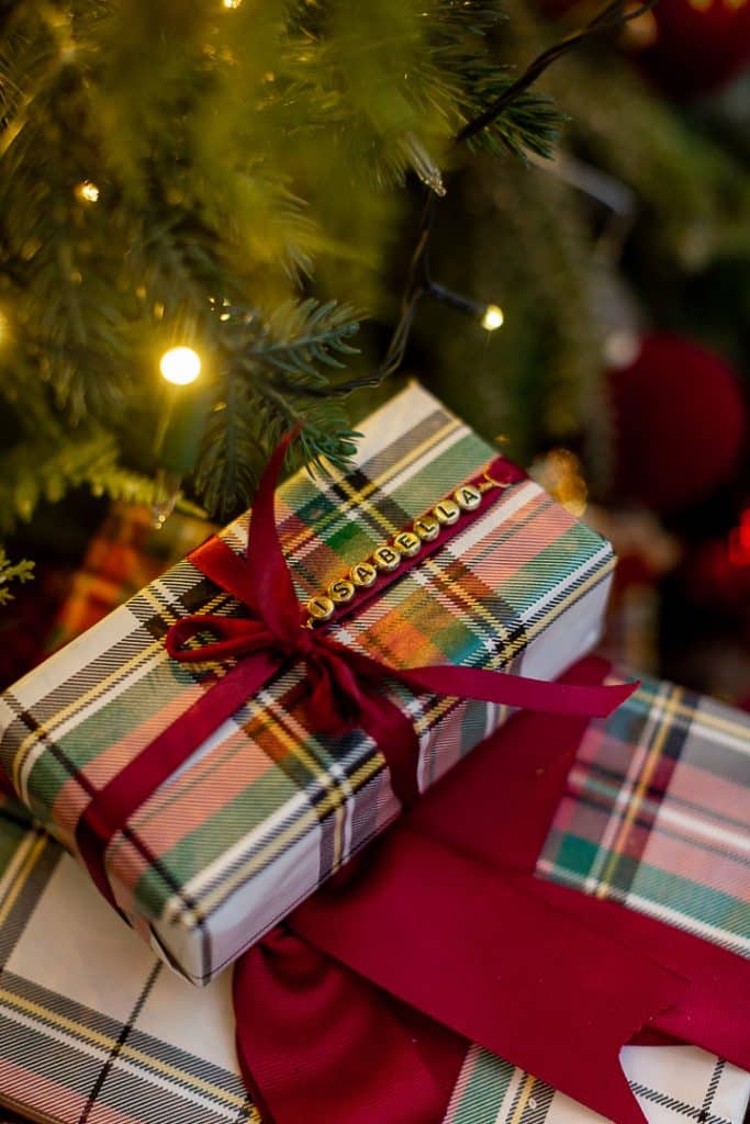 A plaid gift box with a red ribbon and DIY Christmas Gift Tags spelling "Anastasia" sits under a lit Christmas tree, next to another plaid-wrapped present.