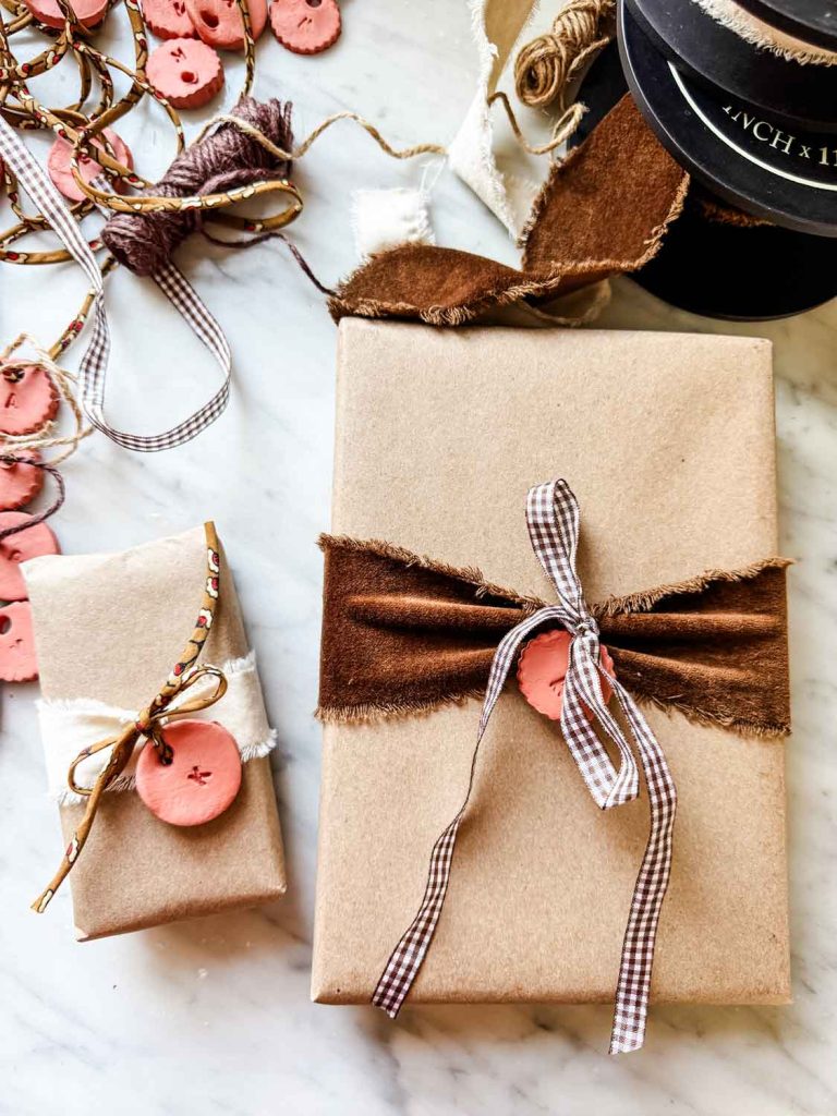 Two gifts wrapped in brown kraft paper sit on a marble surface. Each, adorned with ribbon, pink clay buttons, and DIY Christmas gift tags, is surrounded by craft supplies, ribbons, and more clay buttons scattered nearby.