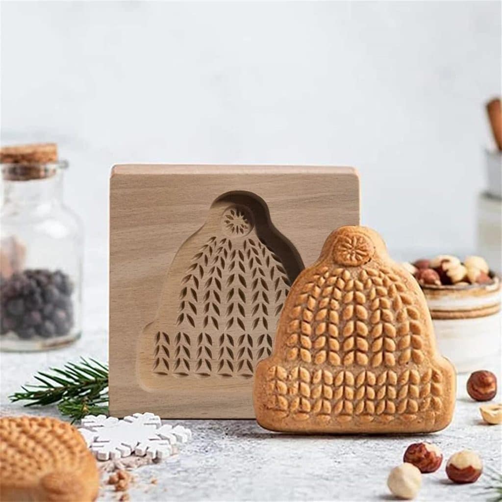 A wooden cookie mold shaped like a knitted winter hat sits on a table next to a molded cookie. Nearby are nuts, spices, and snowflake decorations, creating a cozy scene perfect for trying your favorite Molded Cookies Recipe.