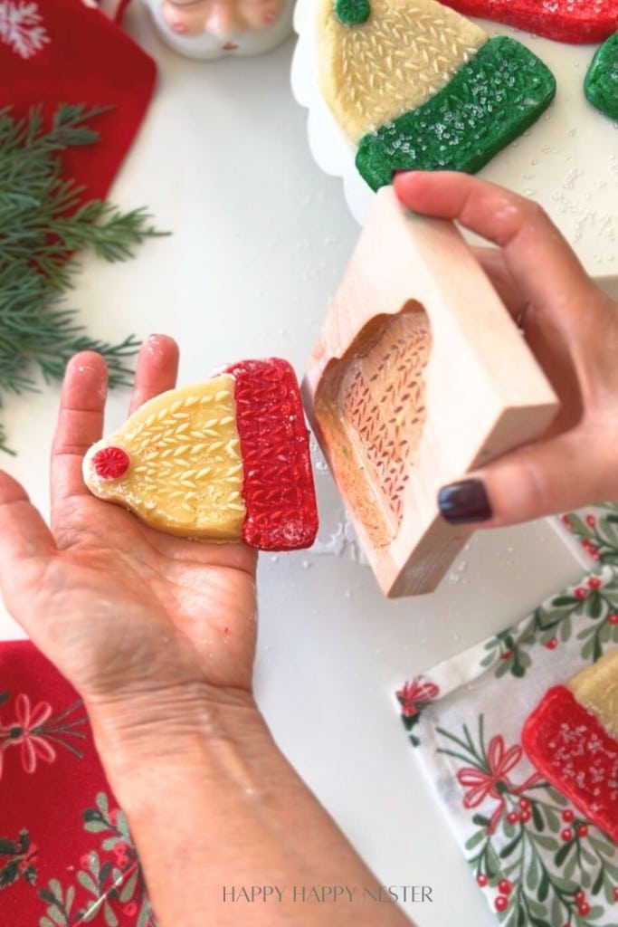A person holds a mitten-shaped holiday cookie decorated with yellow and red icing, just removed from a wooden cookie mold. Decorated napkins and greenery complete the cheerful white table setting.