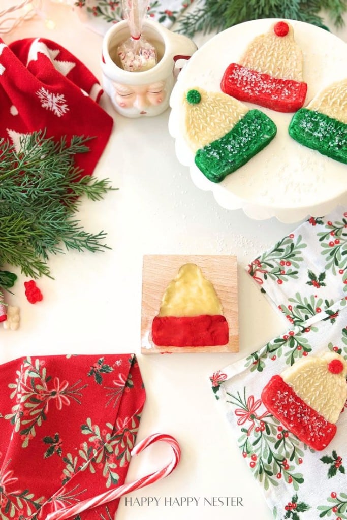 Festive holiday scene with Christmas hats in red and green, a candy cane, greenery, a Santa mug with a spoon, and red and floral napkins on a white surface.