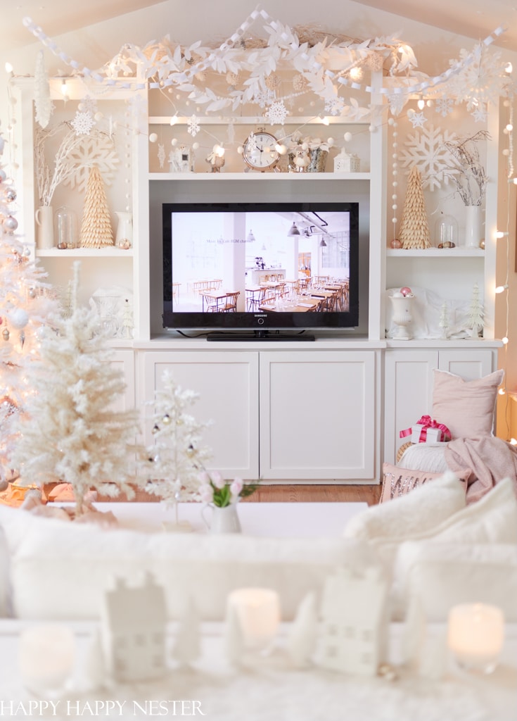 A cozy living room decorated for winter with my favorite Christmas decor—white and gold ornaments, fairy lights, a white Christmas tree, and small houses. The TV is on while gifts, candles, and festive touches fill the bright, airy space.