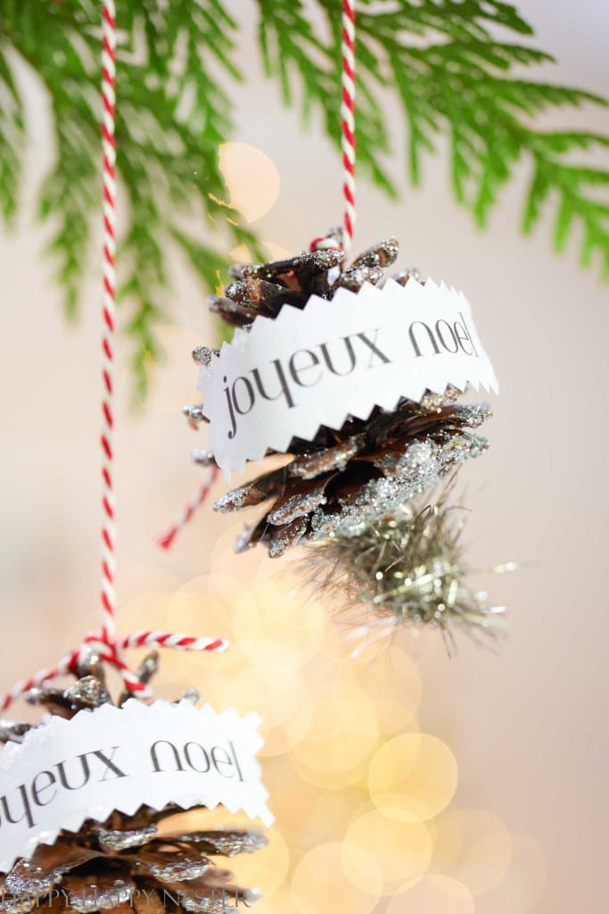 Two pinecone ornaments, my favorite Christmas decor, are decorated with glitter, tinsel, and white paper bands reading "joyeux noel," hanging from red and white string in front of blurred green foliage and warm lights.