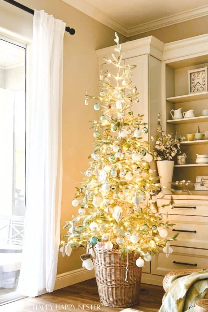 A slim, gold Christmas tree—my favorite Christmas decor—adorned with white and pastel ornaments stands in a wicker basket next to a bright window and cream-colored cabinets in a cozy, light-filled living room.