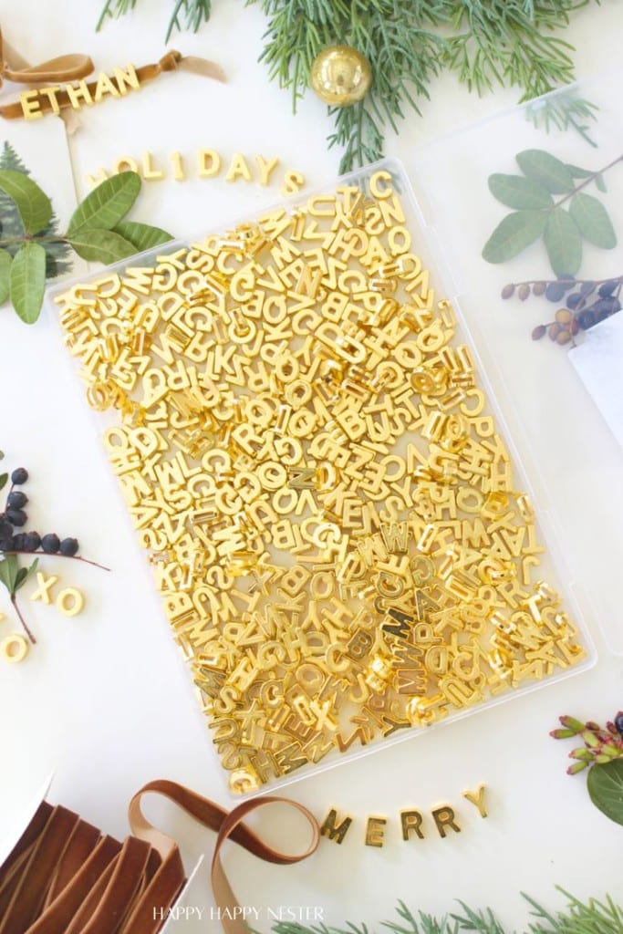 A clear container filled with shiny gold letter charms sits on a white surface, surrounded by green leaves, brown ribbon, holiday decorations, and scattered gold letters spelling “ETHAN,” “HOLIDAY,” “MERRY,” and “XOXO.”.