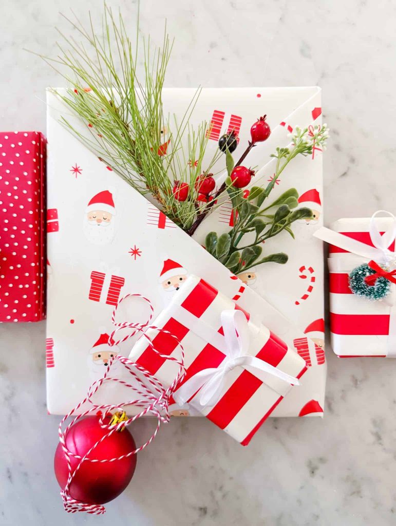 A group of Christmas gifts wrapped in festive paper with red, white, and Santa Claus designs offers my favorite gift wrap inspiration. One is decorated with pine, berries, and mistletoe; a red ornament and white ribbons rest on a marble surface.