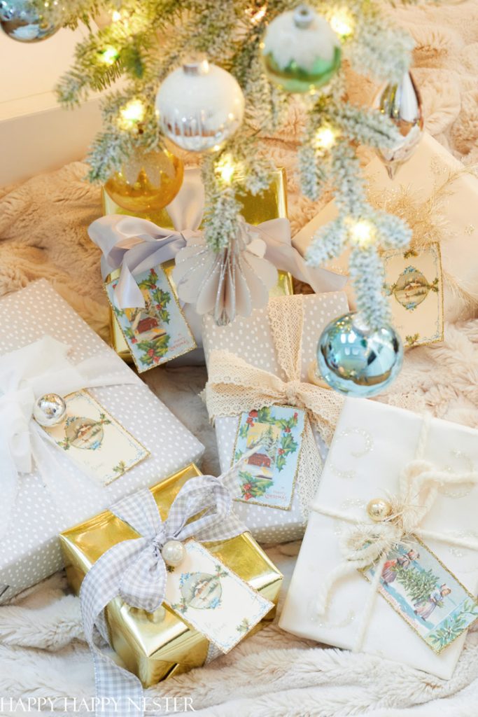 A group of wrapped Christmas gifts with bows and vintage-style tags—my favorite gift wrap inspiration—are arranged under a decorated Christmas tree with white frosted branches and shiny ornaments, all resting on a plush, cream-colored blanket.