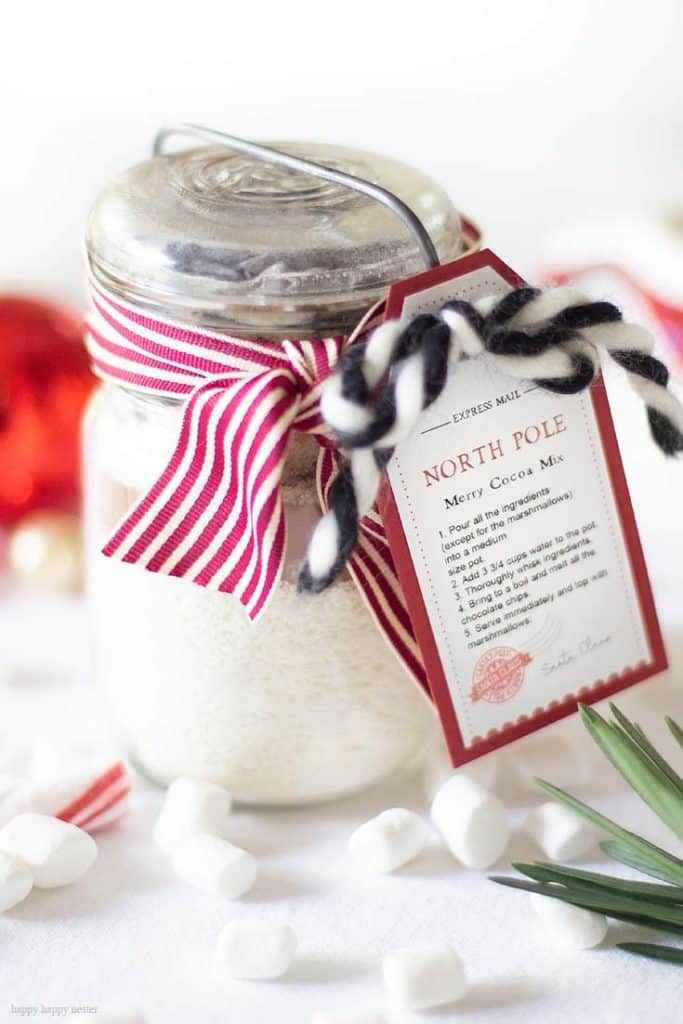 A glass jar filled with cocoa mix is decorated with a red and white striped ribbon, my favorite gift wrap inspiration, and a North Pole-themed tag. Mini marshmallows and holiday decor surround the jar on a white surface.