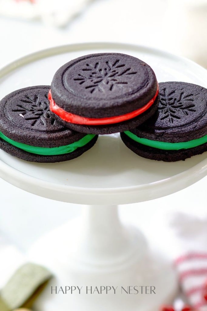 Three chocolate sandwich cookies with red and green cream filling, my favorite Christmas cookies, are displayed on a white cake stand. Each cookie has a festive snowflake design on top.