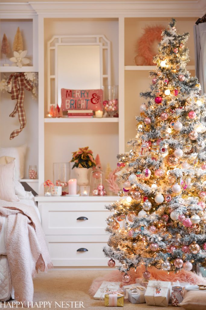 A decorated Christmas tree with pink, gold, and white ornaments—my favorite Christmas decor—stands beside a white bed and a shelf adorned with holiday accents, twinkling lights, and wrapped presents underneath the tree.