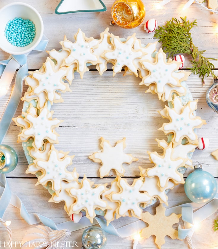 A wreath made of my favorite Christmas cookies—snowflake-shaped sugar cookies with white icing and blue sprinkles—arranged on a white wooden surface with ornaments, ribbon, greenery, and festive lights all around.
