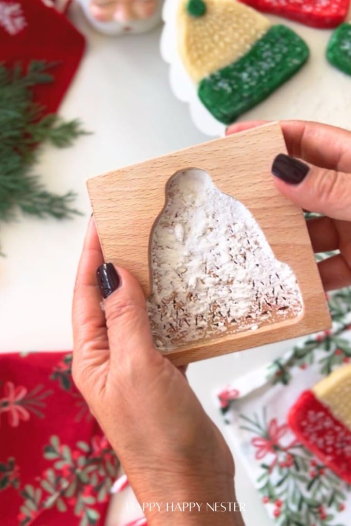 A person with dark nail polish holds a wooden mold filled with powdery white material, shaped like a winter hat—a key step in a molded cookies recipe. Festive cookies and holiday greenery add charm to the background.