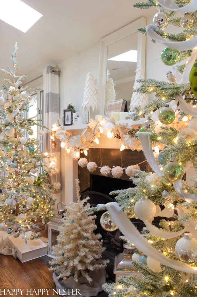 A cozy living room decorated with my favorite Christmas decor—white and silver ornaments, multiple flocked Christmas trees, string lights, and festive garlands on the mantel above a glowing fireplace.