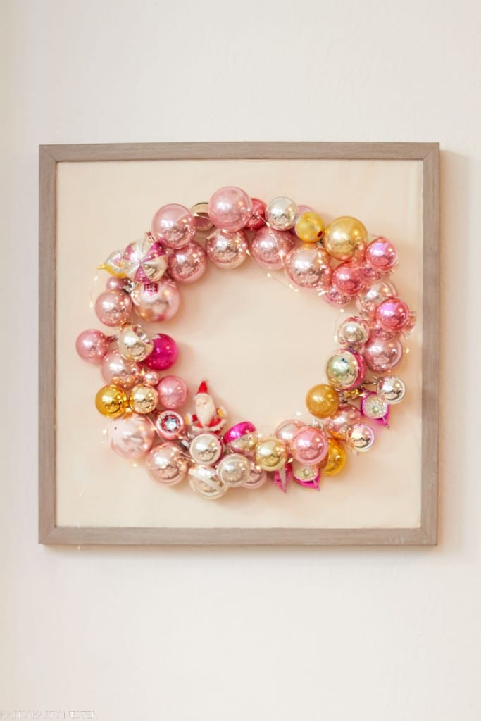 A festive wreath made of pink, gold, and silver ornaments—my favorite Christmas decor—is displayed inside a square, gray-framed shadow box hanging on a light-colored wall.