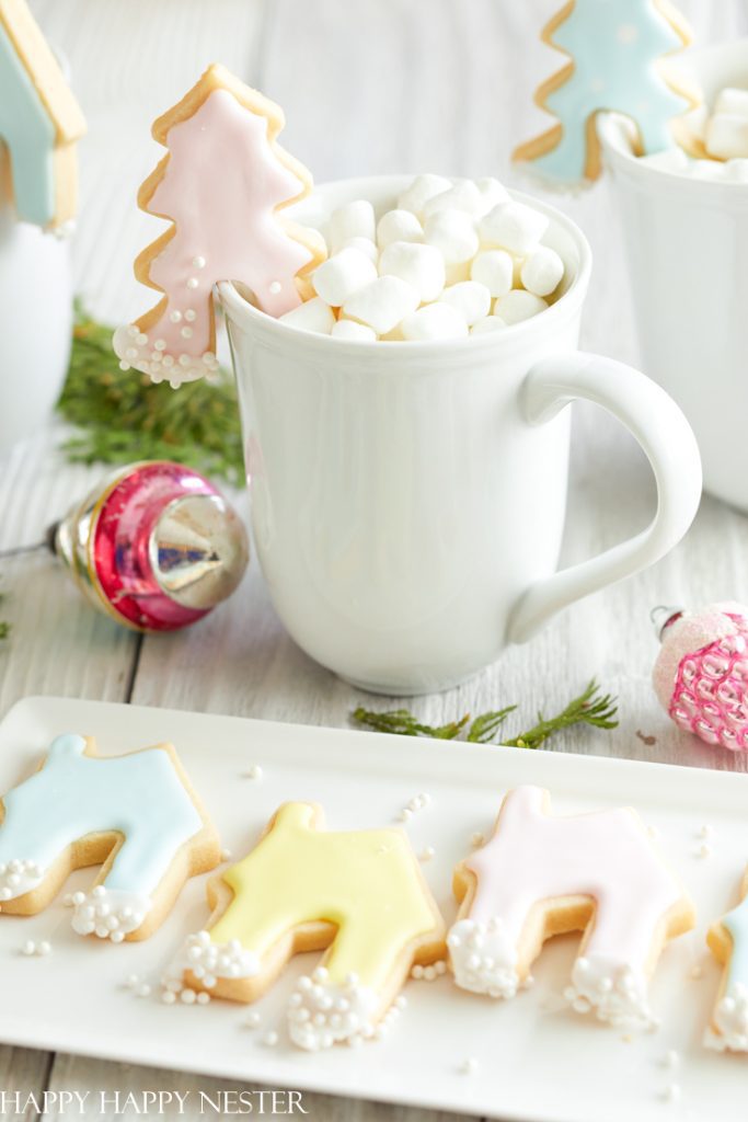 A white mug of hot chocolate with mini marshmallows holds a pastel Christmas tree cookie—one of my favorite Christmas cookies. In front, a plate showcases iced sugar cookies shaped like trees and houses, with ornaments nearby.