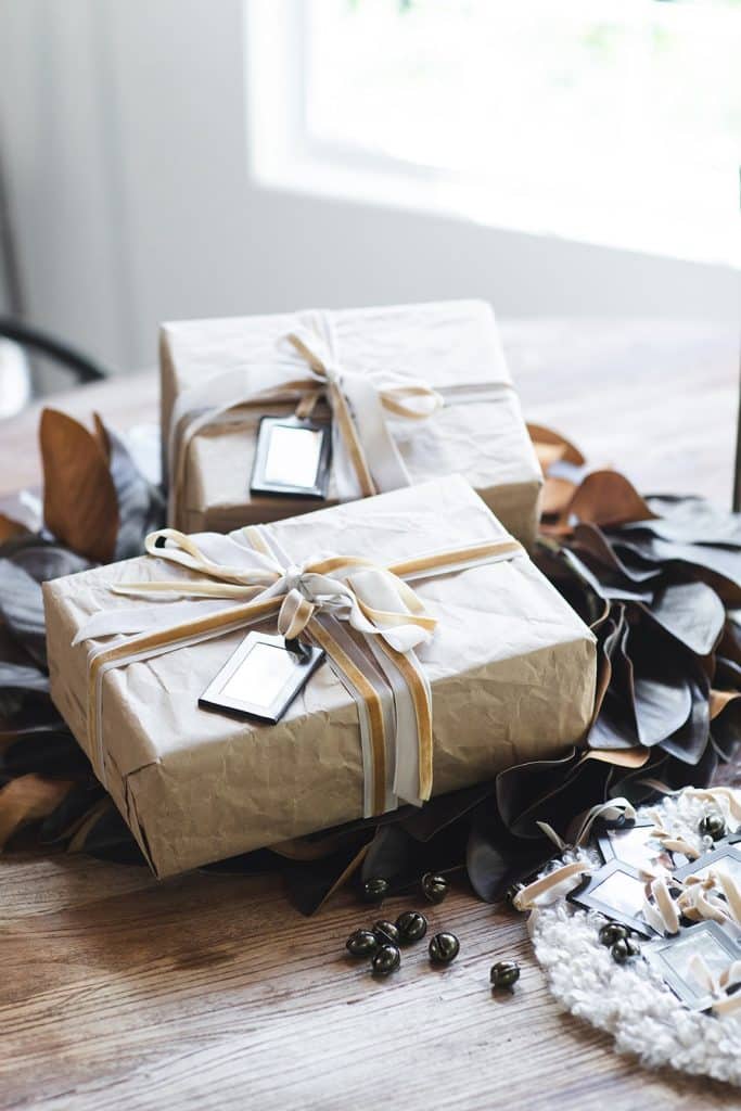 Two gifts wrapped in brown paper and tied with white and gold ribbon, each featuring DIY Christmas Gift Tags, sit on a wooden table decorated with leaves and small ornaments.