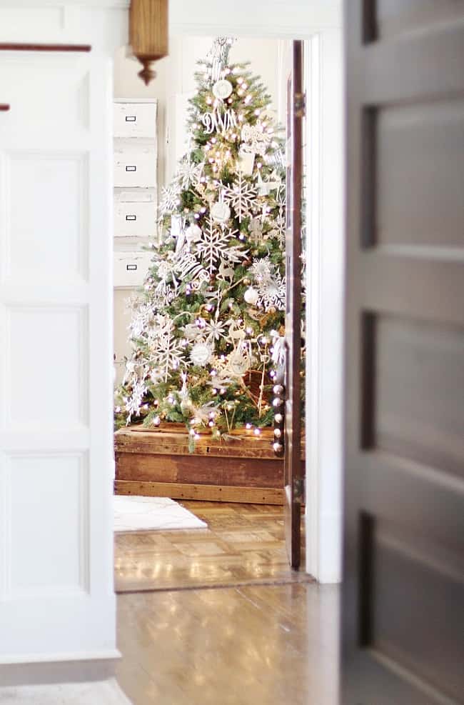 A decorated Christmas tree with white ornaments and lights stands in a bright room, seen through an open doorway. Set in a wooden box, this is my favorite Christmas decor as sunlight reflects off the polished floor.