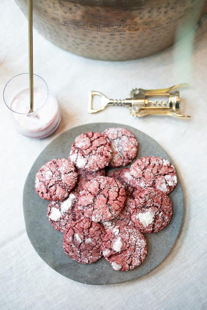 A plate of red crinkle cookies from a classic molded cookies recipe is dusted with powdered sugar, sitting on a table next to a drink with a straw and a metal corkscrew.