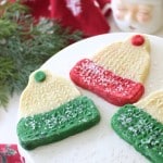 Three holiday cookies shaped like winter hats with red and green bottoms, decorated with white sprinkles, are displayed on a white cake stand with festive greenery and a Santa mug in the background.
