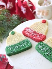 Three holiday cookies shaped like winter hats with red and green bottoms, decorated with white sprinkles, are displayed on a white cake stand with festive greenery and a Santa mug in the background.