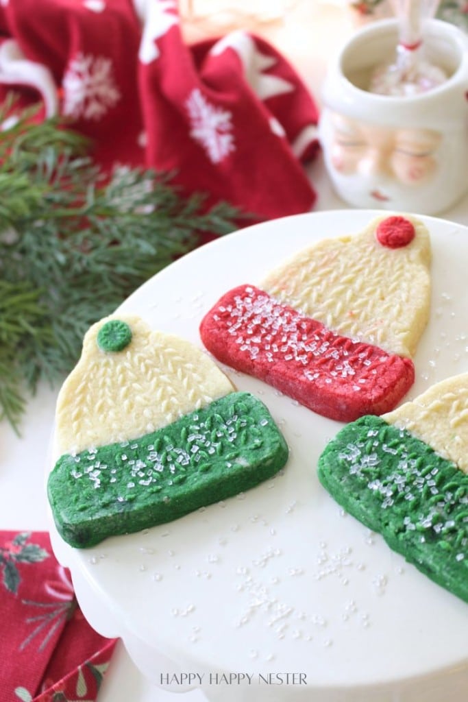 Three holiday cookies shaped like winter hats with red and green bottoms, decorated with white sprinkles, are displayed on a white cake stand with festive greenery and a Santa mug in the background.