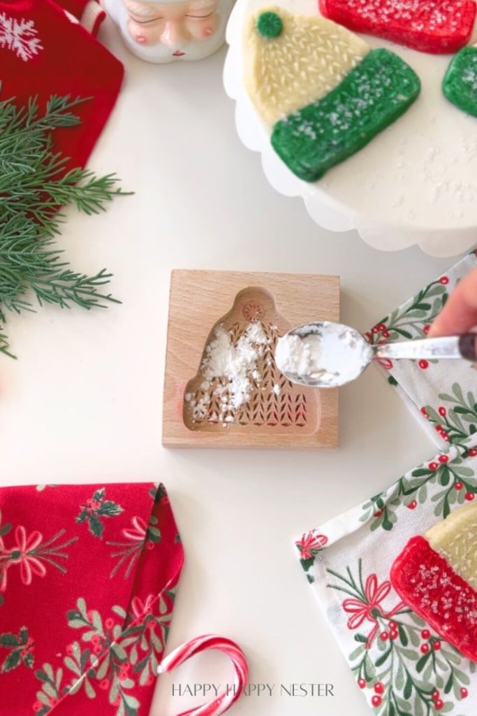 A hand sprinkles powdered sugar into a wooden cookie mold shaped like a mitten, part of a classic molded cookies recipe. Decorated mitten-shaped cookies and festive napkins rest nearby on a white surface with a sprig of greenery.