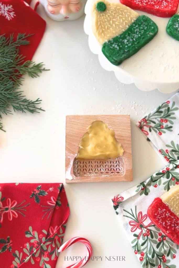 A wooden cookie mold with a Christmas tree design sits on a white surface, surrounded by festive napkins, pine sprigs, decorated cookies made from a classic molded cookies recipe, a candy cane, and a Santa mug.