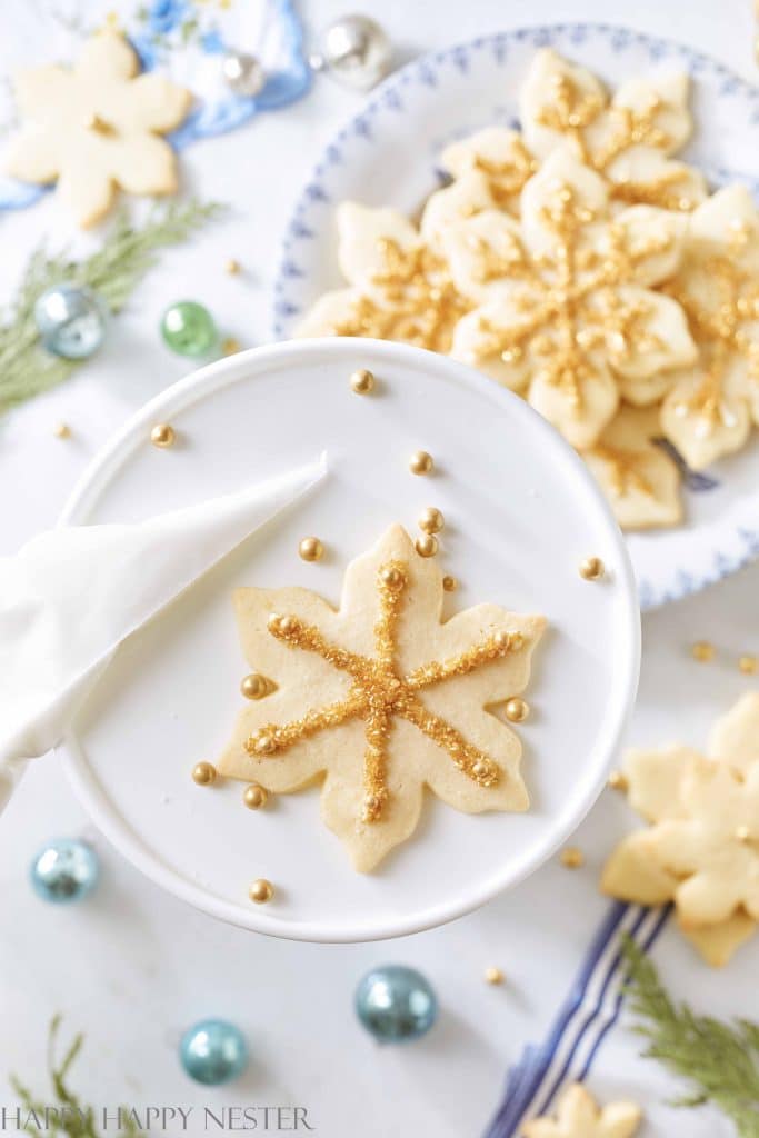 A close-up of one of my favorite Christmas cookies—a leaf-shaped sugar cookie on a white stand, decorated with gold sprinkles and frosting, surrounded by more cookies, festive ornaments, and greenery.