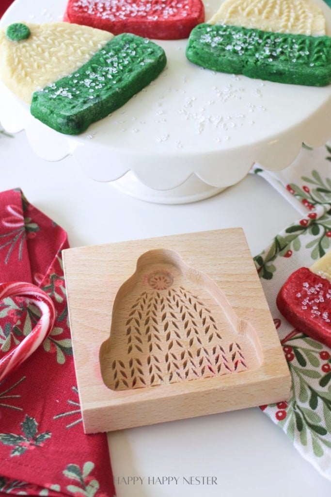 A wooden cookie mold shaped like a winter hat sits on a festive red napkin; decorated red, green, and white molded cookies recipe creations are displayed on a white cake stand in the background.