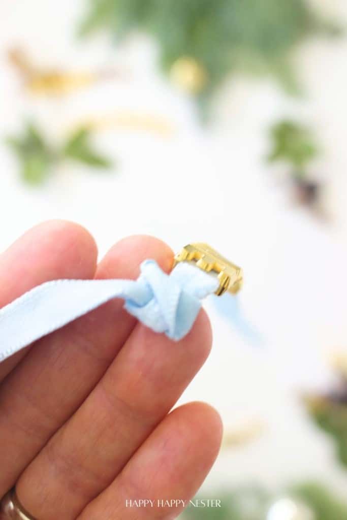 A close-up of a hand holding a light blue ribbon with a gold clasp attached, perfect for diy Christmas gift tags, against a blurred background of greenery and decorations.
