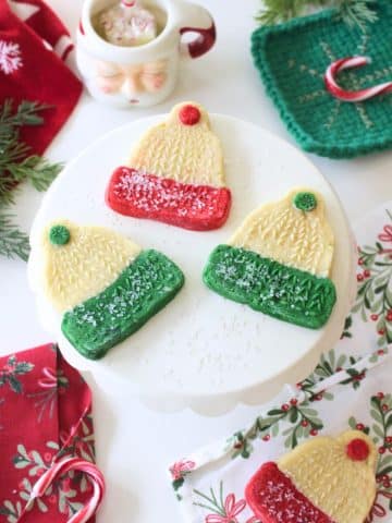 A white cake stand displays four molded cookies shaped like winter hats, decorated with red or green frosting and sprinkles. Festive napkins, pine branches, and a Santa mug surround the display on a white table.