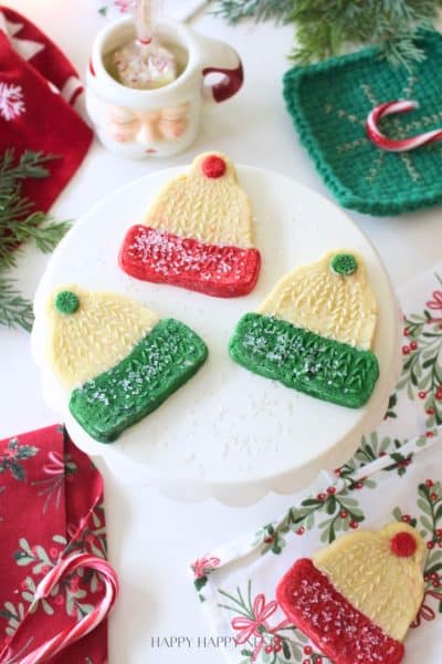A white cake stand displays four molded cookies shaped like winter hats, decorated with red or green frosting and sprinkles. Festive napkins, pine branches, and a Santa mug surround the display on a white table.