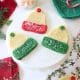 A white cake stand displays four molded cookies shaped like winter hats, decorated with red or green frosting and sprinkles. Festive napkins, pine branches, and a Santa mug surround the display on a white table.
