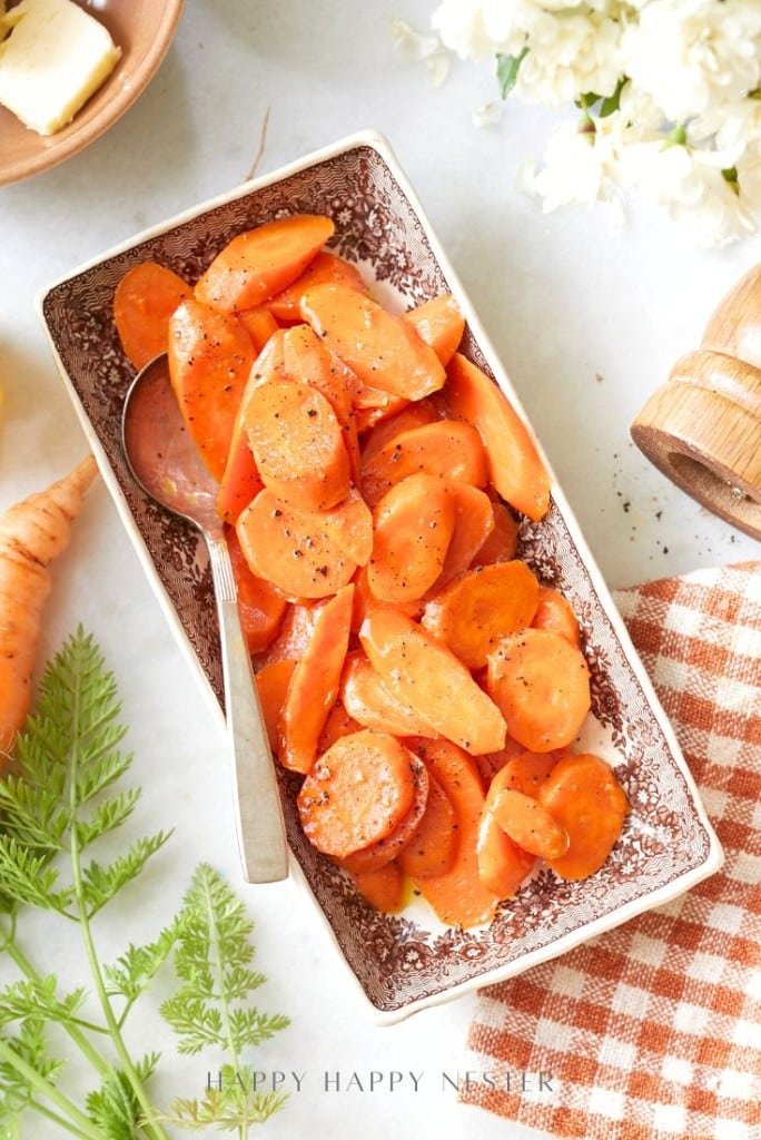 A rectangular dish filled with glazed, sliced carrots garnished with black pepper. Perfect for dinner ideas, this side is served with a spoon and sits beside a checkered cloth, carrot greens, and a wooden pepper grinder.