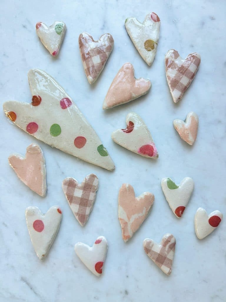 A variety of heart-shaped ceramic pieces are arranged on a white marble surface. Perfect for pink crafts for adults, the hearts are decorated with pastel pinks, polka dots, and plaid patterns in soft, muted colors.