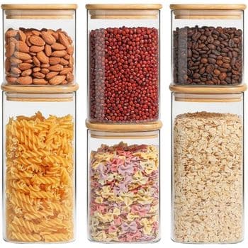 Six glass jars with wooden lids filled with almonds, red beans, coffee beans, pasta spirals, multicolored pasta, and oats, arranged in two rows on a white background.