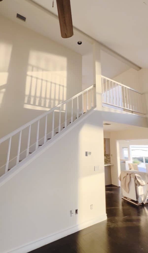 A sunlit, cream-colored interior staircase with white railings leads to an upper loft area—perfect for browsing dinner ideas. Shadows from the railing dance on the wall, while a covered chair sits near a window on the dark floor below.