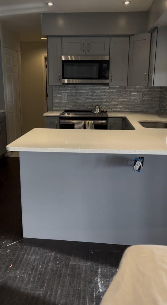 A modern kitchen with gray cabinets, a white countertop, stainless steel appliances, and a tiled backsplash. An outlet without a cover is visible on the island—perfect for powering pink crafts or other creative adult craft ideas. The floor appears unfinished.