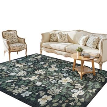 A cream-colored sofa and armchair with floral cushions sit on a dark floral-patterned rug. A round wooden coffee table with a small plant and books is placed in front of the sofa.