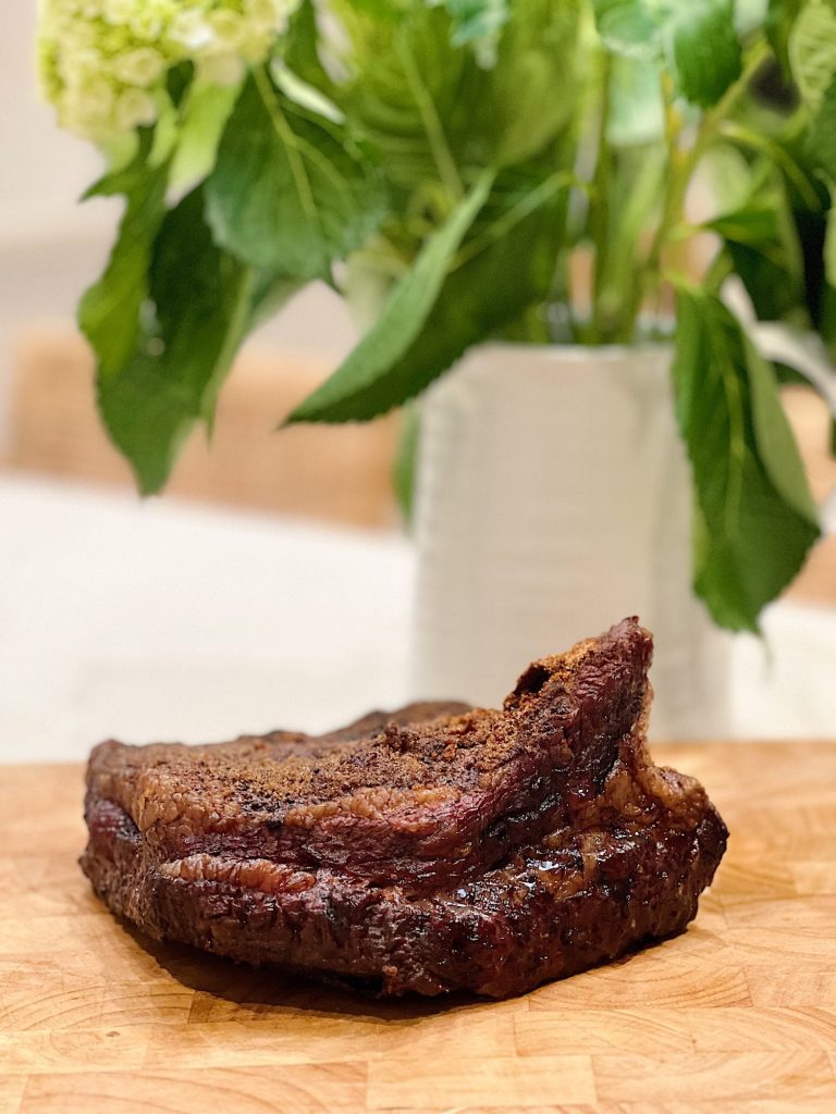 A cooked, seasoned brisket rests on a wooden cutting board with a vase of green leafy flowers blurred in the background, making it perfect for your next dinner ideas.