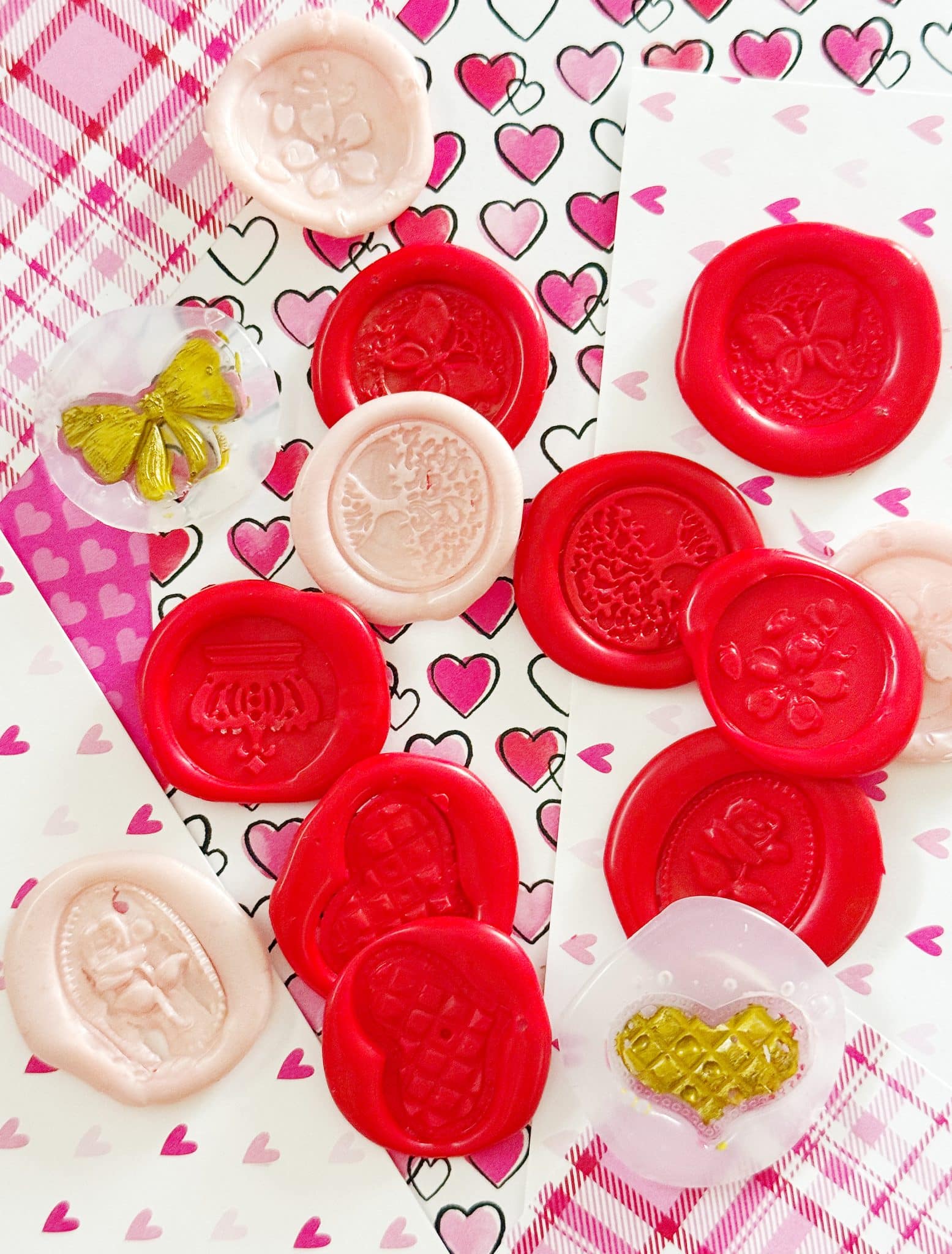 Various pink and red wax seals with intricate designs, including flowers, hearts, and butterflies, are arranged on heart-patterned and plaid paper backgrounds—perfect inspiration for pink DIY projects or crafts for adults.