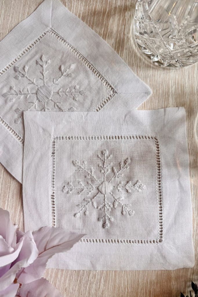 Two white linen napkins with embroidered snowflake designs are displayed on a light wooden surface. A crystal glass and a pale pink flower are visible in the corners of the image.