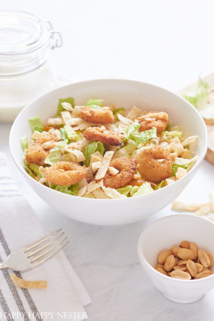 A white bowl filled with salad topped with crispy fried shrimp and tortilla strips, perfect for fresh dinner ideas, with a small bowl of peanuts and a fork nearby on a white table.