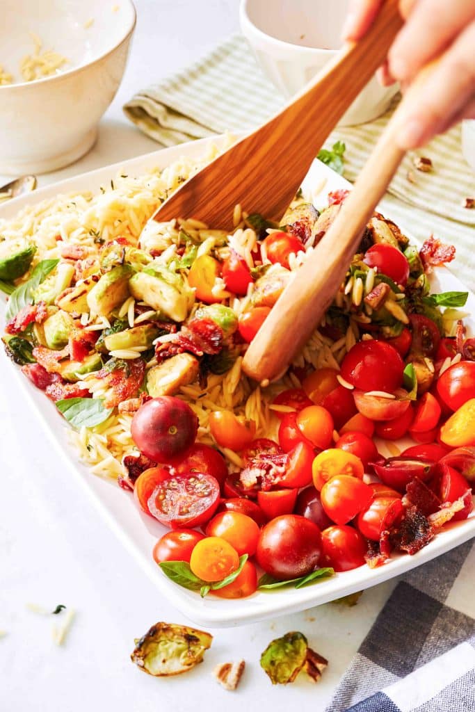 A person tosses a salad with cherry tomatoes, orzo pasta, Brussels sprouts, and bacon in a white bowl using wooden utensils—one of those fresh dinner ideas perfect for any night. In the background are bowls and a striped cloth.