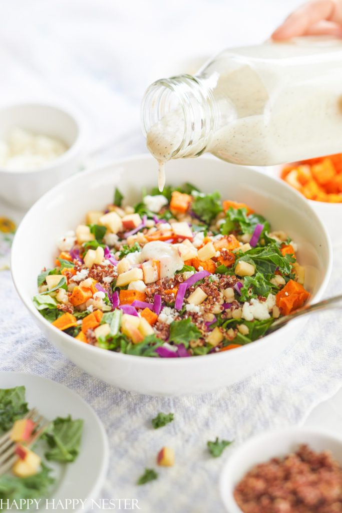 A hand pours creamy dressing from a glass bottle over a colorful salad with kale, quinoa, apples, sweet potatoes, and red onions in a white bowl—perfect for fresh and healthy dinner ideas. Small bowls of salad ingredients are visible in the background.